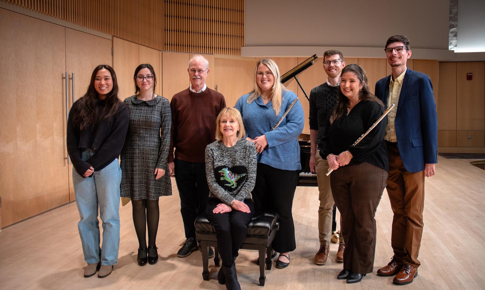 Group of eight people gathers on stage next to piano for a photo