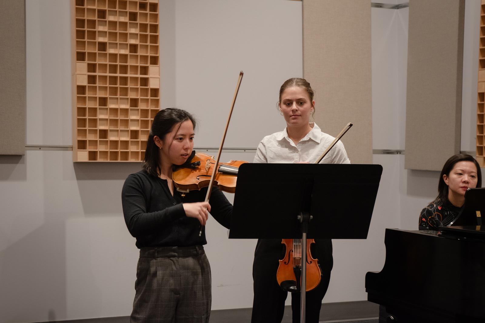Person playing violin and instructing a student while someone plays piano in a classroom