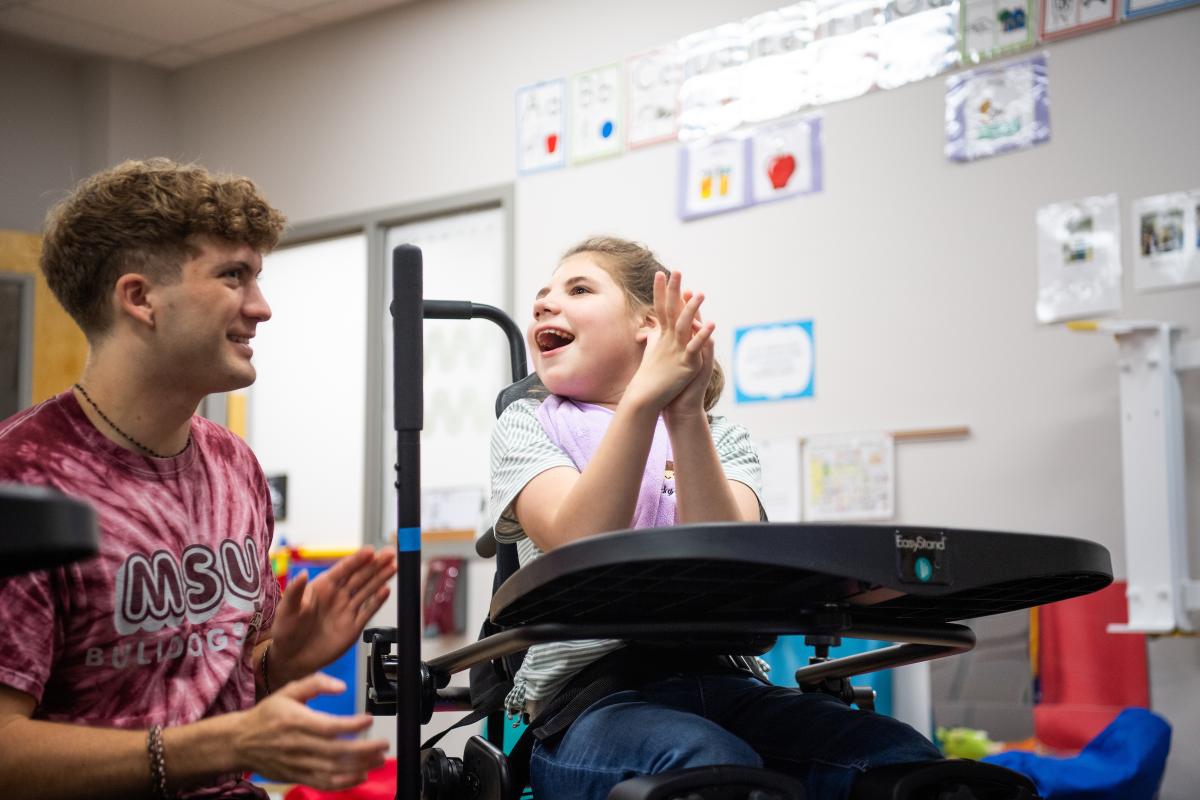Staff member sitting with child in a wheelchair, clapping hands and smiling